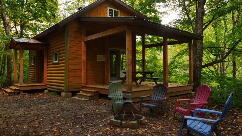 Façade du refuge Le Draveur, avec à l'avant-plan l'espace détente extérieur avec ses chaises et son foyer extérieur, entouré par la nature verdoyante de la Vallée, l'un des hébergements offerts dans la gamme des Refuges, en pleine nature.