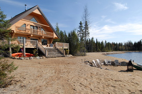 View of the front of one of the chalets offered by Au Chalet en Bois Rond, a Partner nature village in La Vallée, with its sand-covered beach and lake surrounded by nature.